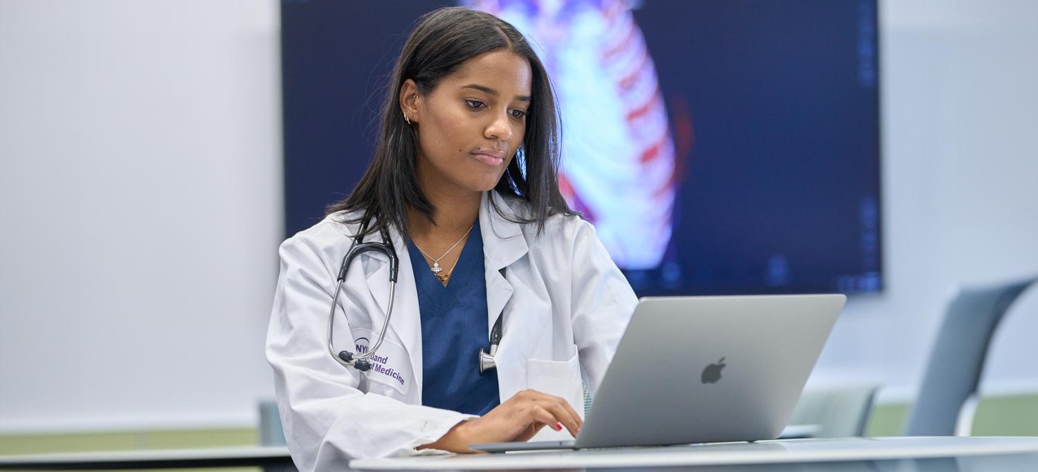 Resident wearing a white coat and stethoscope around shoulders typing on a laptop computer