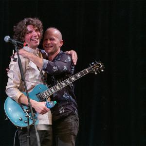 Jesse Kinch, Wearing Guitar on Strap, and Dr. Todd J. Carpenter Embrace on Stage During Performance, While Seated Bandmate Looks On